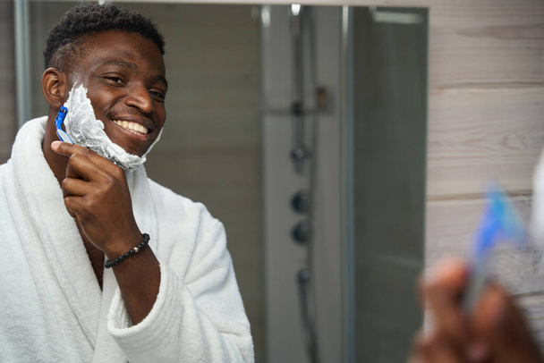 African American guy shaves in front of the mirror in the bathroom, he is wearing a terry robe - Photo, Image