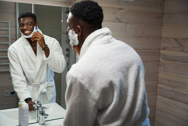 Afrikai amerikai férfi borotválkozik előtt egy nagy tükör a fürdőszobában, visel frottír köntös - Fotó, kép