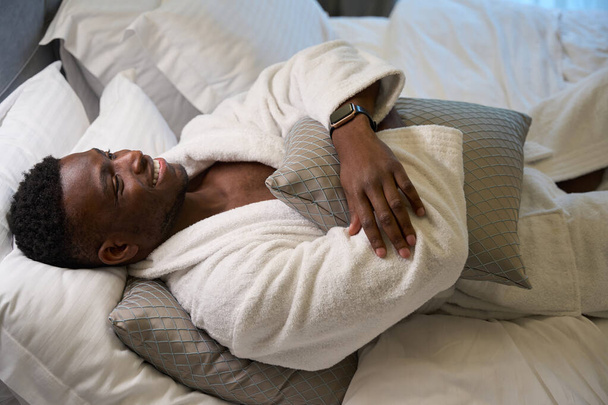 Guy geniet van 's morgens ontspanning in een gezellige slaapkamer, hij is in een zachte badjas - Foto, afbeelding