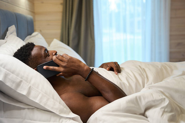 Homem de cabelos encaracolados se comunica em um telefone celular na cama, ele se encontra sob um cobertor macio - Foto, Imagem
