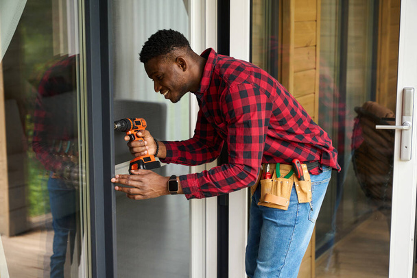 Αφροαμερικάνος επισκευαστής επισκευάζει μια γυάλινη πόρτα, χρησιμοποιεί ένα κατσαβίδι - Φωτογραφία, εικόνα