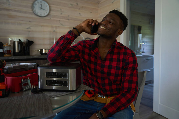 Glimlachende reparateur communiceert op een mobiele telefoon, hij doet kleine reparaties in de keuken - Foto, afbeelding