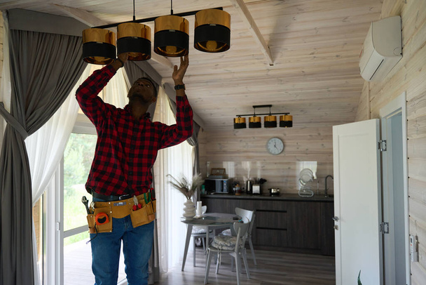 Afroamerikaner repariert einen Kronleuchter in der Küche, er hat einen Werkzeuggürtel - Foto, Bild