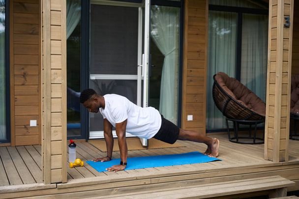 African American guy doing fitness on the terrace of an eco-cottage, next to dumbbells and a bottle of water - Photo, Image