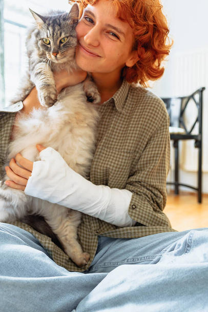teen girl with curly red hair, broken arm in cast, sits on floor of house, hugging favorite fluffy cat. Friendship teenager pet - Fotó, kép