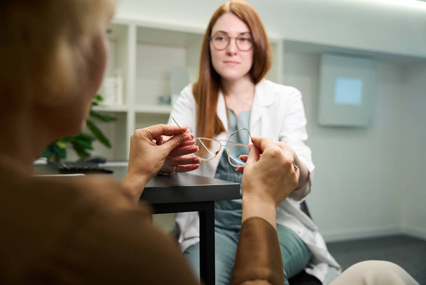 Σύμβουλος φροντίδας συνιστά ένα κατάλληλο πλαίσιο για τα γυαλιά σε έναν πελάτη, κυρίες επικοινωνούν σε ένα σύγχρονο σαλόνι οπτικής - Φωτογραφία, εικόνα