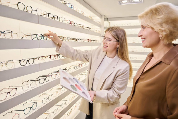 Consultor oftalmologista em um salão óptico oferece uma moldura adequada para um cliente, existem muitos quadros diferentes em exibição - Foto, Imagem
