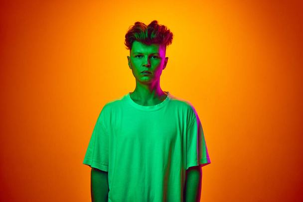 Portrait de jeune homme calme, gars, étudiant en t-shirt blanc isolé sur fond dégradé orange illuminé lumière au néon vert. Concept de jeunesse, beauté, mode, tendances émotions, ventes. publicité - Photo, image