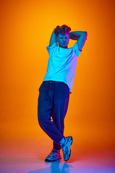 Full length portret van jonge aantrekkelijke man zet zijn handen achter het hoofd gekleed terloops weg te kijken tegen gradiënt oranje achtergrond verlicht neon licht. Concept van jeugd, schoonheid, mode, stijl - Foto, afbeelding