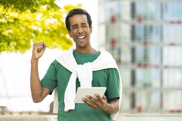 Портрет улыбающегося позитивного афроамериканца, держащего в руках цифровой планшет, получающего хорошие новости, делающего жест победителя, работающего онлайн в парке. Концепция успешного бизнеса - Фото, изображение
