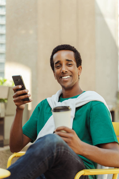 Muotokuva hymyilevä positiivinen Afrikkalainen Amerikkalainen mies istuu kahvilassa pitäen matkapuhelin ja kahvikuppi juomalla, rentouttava, katsoen kameraa. Kahvitauon käsite - Valokuva, kuva