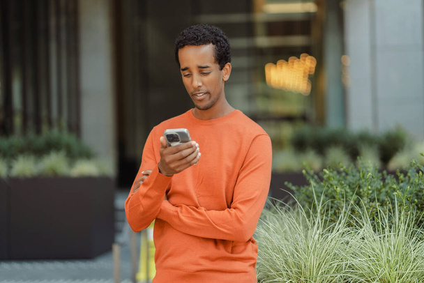 Retrato de atraente sorridente afro-americano homem segurando telefone celular leitura mensagem de texto, comunicação on-line na rua  - Foto, Imagem