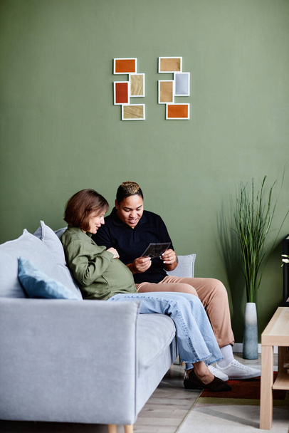 Minimal shot of modern gay couple with pregnant young woman looking at pictures of baby in home interior against green wall, copy space - Photo, image