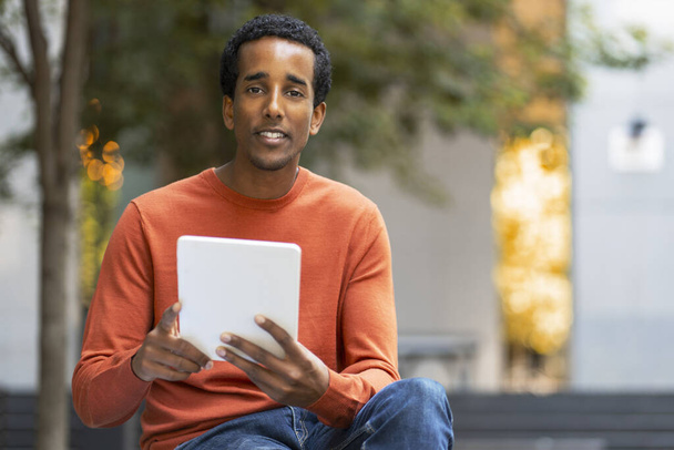 Porträt eines lächelnden Afroamerikaners, der ein digitales Tablet in der Hand hält, E-Books liest, auf der Straße sitzt, im Freien Raum kopiert. Schöner Geschäftsmann, der E-Mails und SMS checkt. Technologiekonzept - Foto, Bild