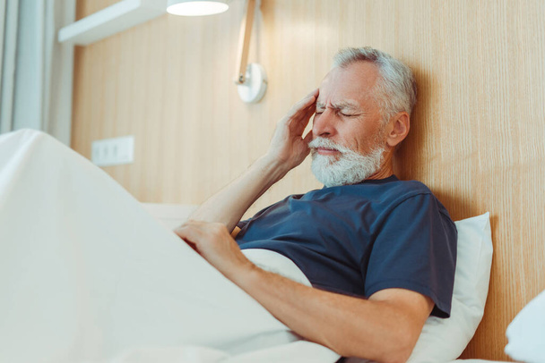 Triste uomo anziano stanco che ha mal di testa, emicrania, sentirsi male sdraiato nel letto d'ospedale. Dolore, stress, concetto di trattamento medico  - Foto, immagini