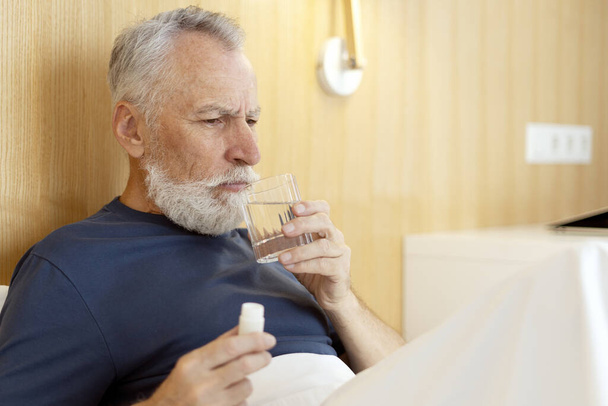 Zdenerwowany, zmęczony starszy mężczyzna leżący w łóżku, trzymający słoik z lekarstwami i szklankę wody, odwracając wzrok. Emeryt bierze tabletki, ma problemy ze zdrowiem. Koncepcja leczenia - Zdjęcie, obraz