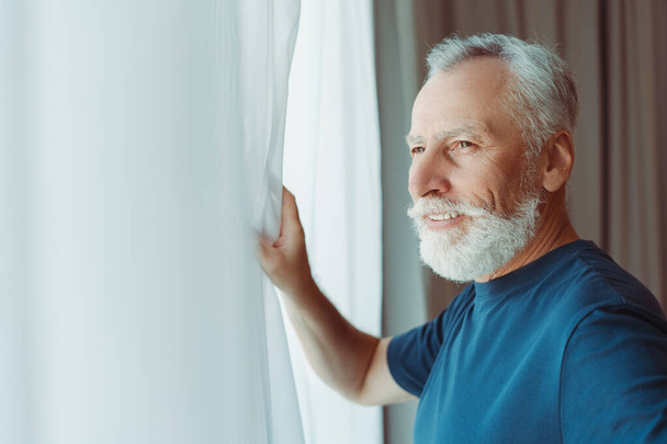 Ritratto di uomo anziano barbuto sorridente che indossa una t-shirt blu guardando la finestra, copiare spazio. Concetto mattina  - Foto, immagini