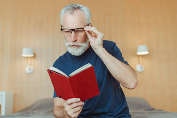 Portrét zamyšleného staršího muže, který nosí brýle, drží zápisník, čte knihu, sedí na posteli v obývacím pokoji. Starší důchodce v neformálních šatech, studuje. Koncept vzdělávání - Fotografie, Obrázek