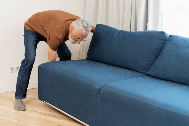 Πορτρέτο του ηλικιωμένου με γυαλιά κινείται καναπέ στο διαμέρισμα. Ελκυστικός ηλικιωμένος συνταξιούχος στο σαλόνι. Έννοια του εσωτερικού - Φωτογραφία, εικόνα