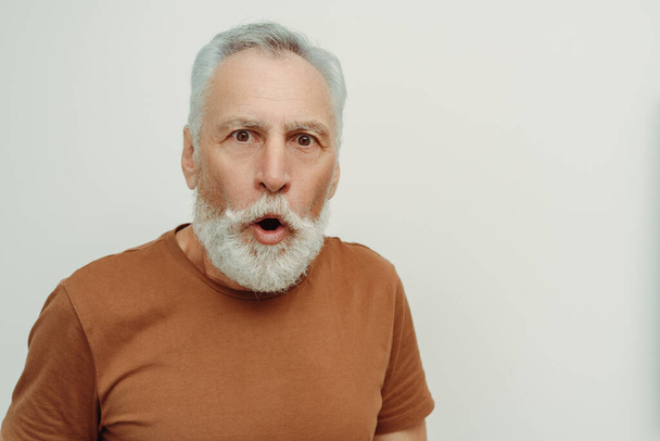 Amazed, shocked senior man wearing brown t shirt looking at camera isolated on gray background. Excited male posing for picture, copy space. Concept of shopping, sale - Photo, Image