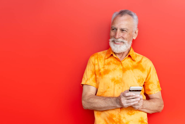 Красивий усміхнений старший чоловік тримає покупки мобільних телефонів онлайн на вулиці ізольовано на червоному тлі, копіює простір. Бородатий хіпстер використовує смартфон, спілкування онлайн. Мобільний банкінг  - Фото, зображення