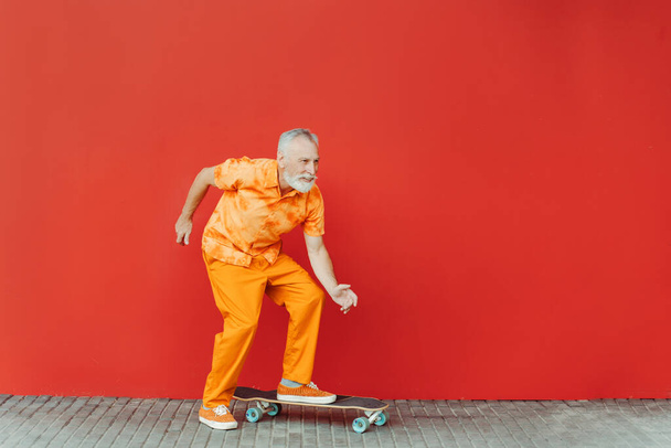 Hombre mayor elegante guapo, hipster barbudo con ropa casual naranja montando monopatín en la calle, cerca de la pared roja. Publicidad, concepto de estilo de vida positivo  - Foto, imagen