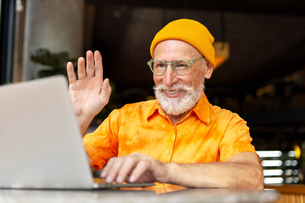 Hombre mayor guapo, hipster sonriente usando computadora portátil saludando la mano teniendo videollamada sentada en la cafetería moderna. Tecnología, concepto de videoconferencia  - Foto, Imagen