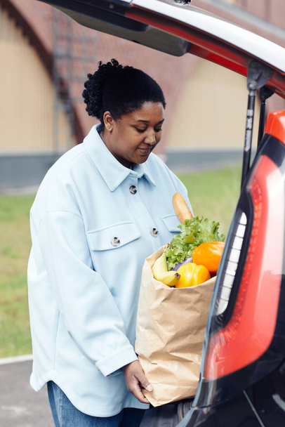 Pystysuora muotokuva nuori musta nainen laittaa ruokakassi auton takakonttiin supermarket parkkipaikalla - Valokuva, kuva
