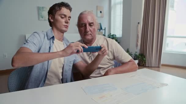 Средний снимок маленького сына, обучающего своего отца на пенсии использовать приложение для оплаты на смартфоне, сидя за столом - Кадры, видео