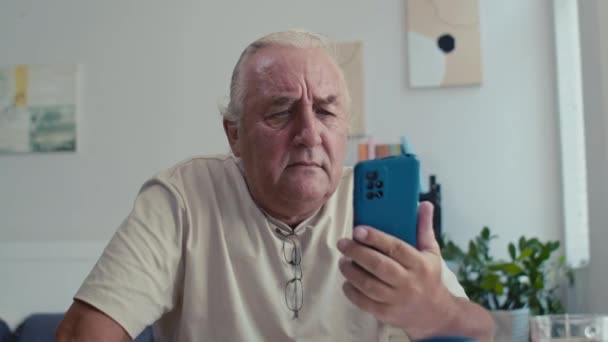 Średnie ujęcie smutnego starszego mężczyzny mającego problemy podczas korzystania z nowoczesnego smartfona w domu - Materiał filmowy, wideo
