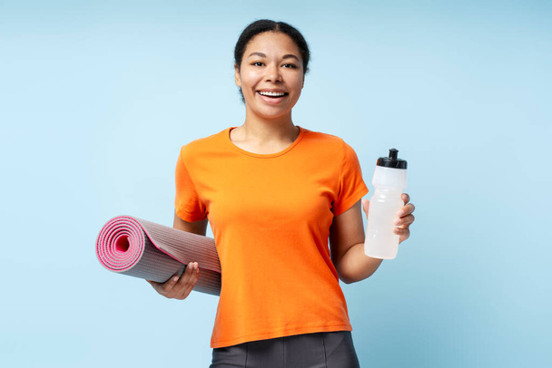 Afroameričanka šťastný úsměv žena drží fitness jóga podložka a láhev vody, izolované na modrém pozadí  - Fotografie, Obrázek