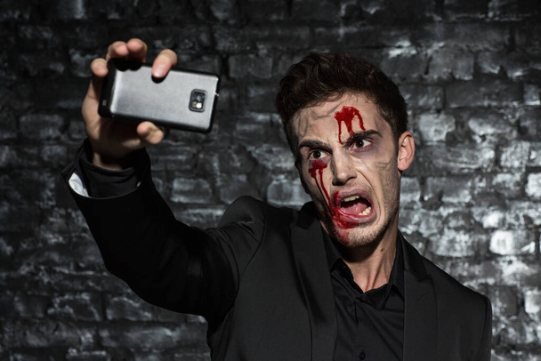 Egy fiatal férfi portréja, fekete öltönyben, véres sminkkel Halloweenkor, amint szelfit csinál az okostelefonján ijesztő arccal. - Fotó, kép