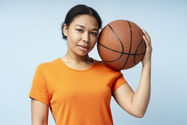 Портрет красивой улыбающейся афроамериканки, играющей в баскетбол, держащей мяч, смотрящий в камеру, изолированный на синем фоне. Спорт, хобби, концепция здорового образа жизни - Фото, изображение