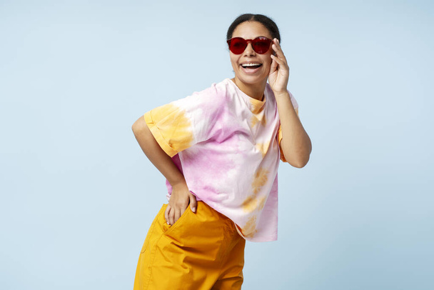 Portrait of beautiful smiling African American woman wearing trendy tie dye t shirt and sunglasses isolated on blue background. Smiling stylish fashion model having fun posing for picture in studio  - Photo, Image