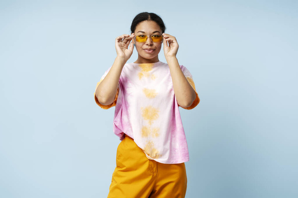 Portrait of beautiful confident African American woman wearing trendy tie dye t shirt, stylish sunglasses isolated on blue background. Attractive stylish fashion model posing for picture in studio - Photo, Image
