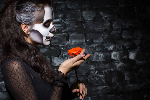 Trieste jonge vrouw met skelet make-up op het gezicht te kijken naar oranje bloem in de studio. Zijaanzicht van attente vrouw met decoratieve Halloween make-up, met gerbera, kopieerruimte. Halloween concept.  - Foto, afbeelding