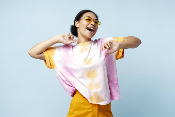 Smiling African American woman dancing wearing trendy t shirt and eyeglasses isolated on blue background. Happy stylish fashion model having fun posing for picture in studio - Photo, Image