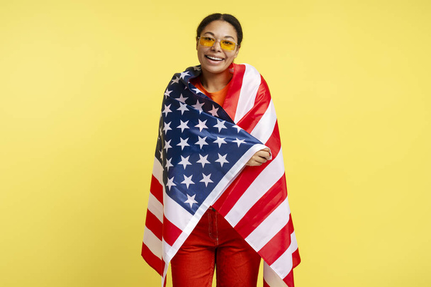 Портрет уверенно улыбающейся афроамериканки с американским флагом на желтом фоне, отмечающей День независимости. 4 июля  - Фото, изображение