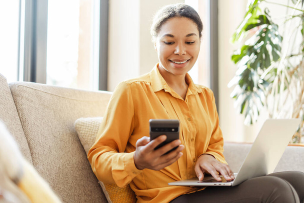 Moderna mulher de negócios afro-americana sorridente, freelancer segurando telefone celular, usando laptop trabalhando online em casa. Elegante redator com smartphone sentado no café. Negócios bem sucedidos - Foto, Imagem