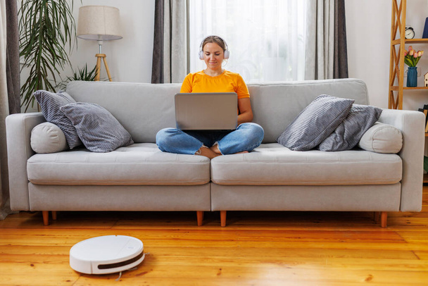 Robotstofzuiger schoonmaken van de woonkamer terwijl de vrouw met behulp van laptop zittend op de bank thuis - Foto, afbeelding