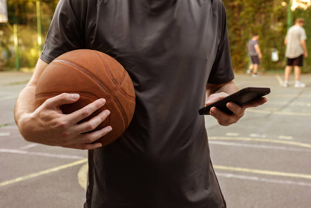 A man with a basketball ball and a phone on an open sports court. Sports concept. - Foto, imagen