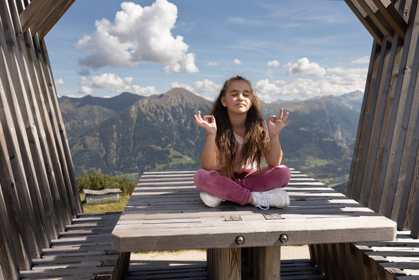 Menina bonito de cabelos longos sentado na mesa de madeira em gazebo na posição de lótus no fundo das montanhas e do céu. Foto de alta qualidade - Foto, Imagem