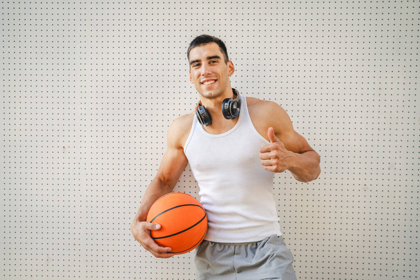 Egy fiatal kaukázusi férfi férfi sportoló állni kültéri hold kosárlabda viselet fehér tartály felső ing erős izmos valós személy másolja tér boldog mosoly magabiztos egészséges életmód koncepció - Fotó, kép