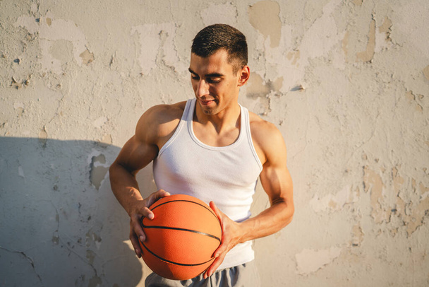 Egy fiatal kaukázusi férfi férfi sportoló állni kültéri hold kosárlabda viselet fehér tartály felső ing erős izmos valós személy másolja tér boldog mosoly magabiztos egészséges életmód koncepció - Fotó, kép