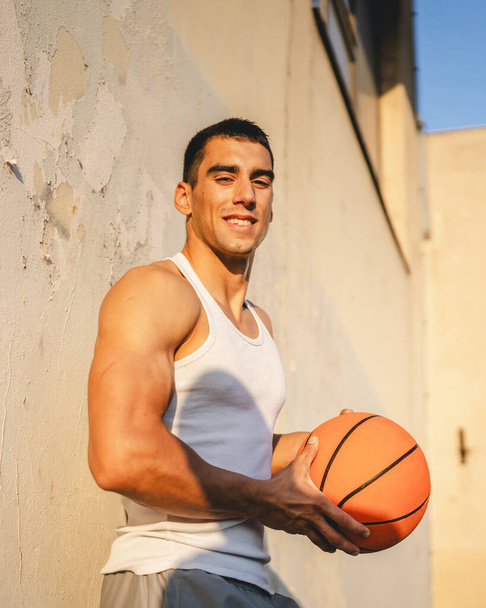 Ένας νεαρός καυκάσιος άνδρας άνδρας αθλητής σταθεί υπαίθρια κρατήστε μπάλα μπάσκετ φορούν λευκό δεξαμενή μπλούζα ισχυρή μυϊκή πραγματικό πρόσωπο αντίγραφο χώρο χαρούμενο χαμόγελο αυτοπεποίθηση υγιή τρόπο ζωής έννοια - Φωτογραφία, εικόνα