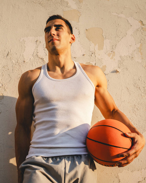 jeden młody kaukaski mężczyzna mężczyzna atleta stoją na zewnątrz trzymać koszykówka piłka nosić biały zbiornik top a-shirt silny umięśniony prawdziwa osoba kopia przestrzeń szczęśliwy uśmiech pewny zdrowy styl życia koncepcja - Zdjęcie, obraz