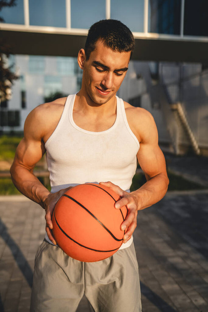 Один молодой кавказский мужчина спортсмен стоять на открытом воздухе провести баскетбольный мяч носить белый топ танк футболка сильный мускулистый реальный человек скопировать пространство счастливая улыбка уверенно здоровый образ жизни - Фото, изображение