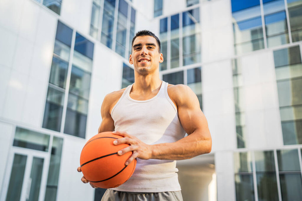 Een jonge blanke man mannelijke atleet staan outdoor hold basketbal bal dragen witte tank top een-shirt sterk gespierd echte persoon kopiëren ruimte gelukkig glimlach zelfverzekerd gezonde levensstijl concept - Foto, afbeelding