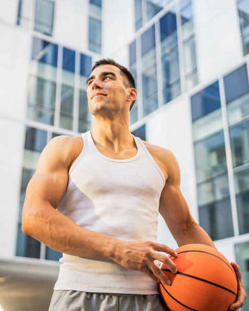 Een jonge blanke man mannelijke atleet staan outdoor hold basketbal bal dragen witte tank top een-shirt sterk gespierd echte persoon kopiëren ruimte gelukkig glimlach zelfverzekerd gezonde levensstijl concept - Foto, afbeelding
