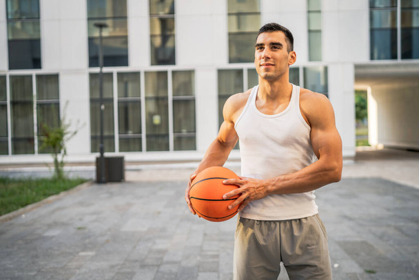 Один молодий білий чоловік чоловік, спортсмен, стоїть на відкритому повітрі, тримає баскетбольну м'яч, одягнений у білий бак, зверху футболка сильна м'язова реальна людина копіює простір щасливої посмішки впевнена концепція здорового способу життя - Фото, зображення